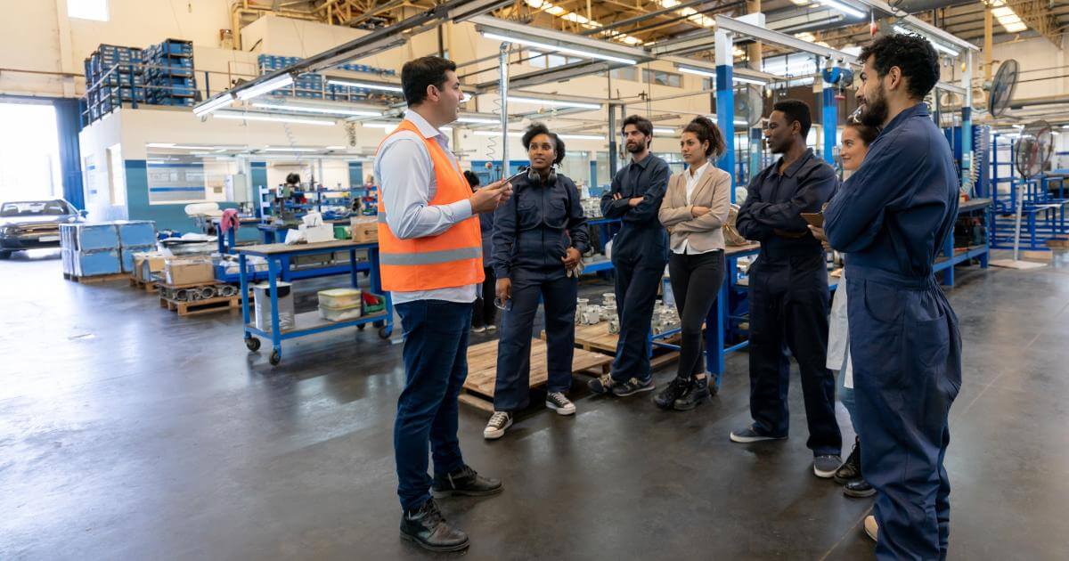 Manufacturing supervisor in a safety vest addressing a group of workers on the production floor