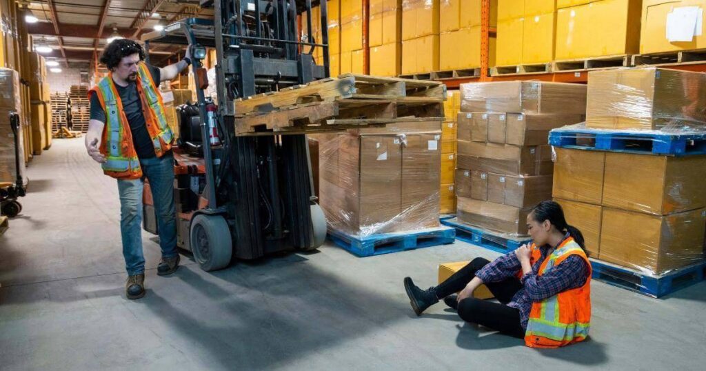 warehouse worker seated on the floor after an accident near a forklift while a colleague looks on