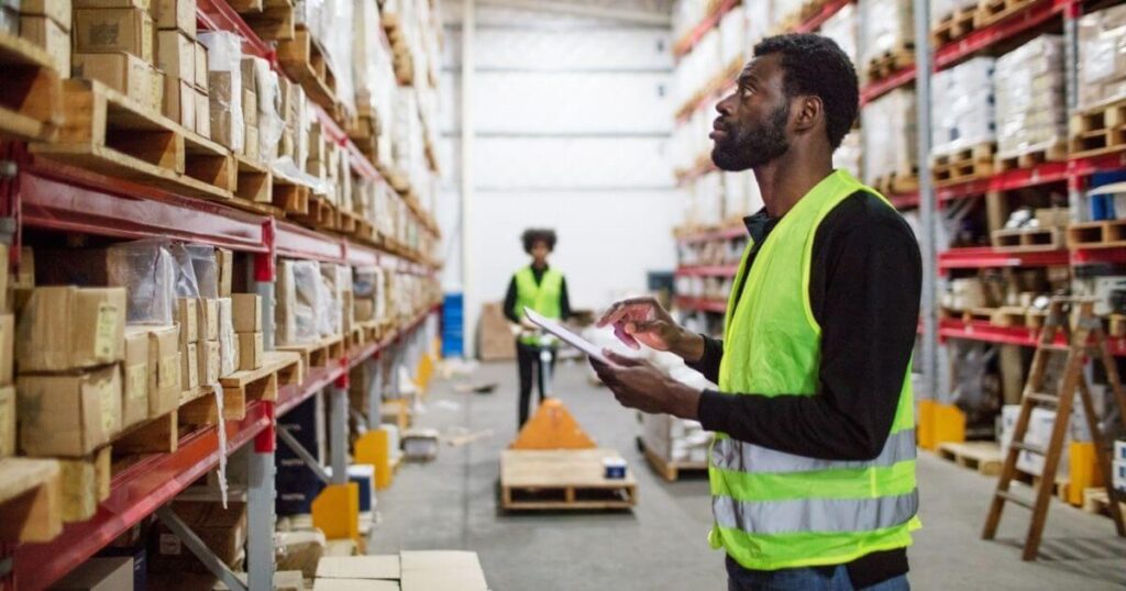 Warehouse worker in a high-visibility safety vest checking inventory with a clipboard while looking up at pallet racking, with a second worker in the background